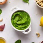 Top down image of spinach cashew pesto in a ceramic bowl on a light gray background with ingredients poking into the edges of the frame