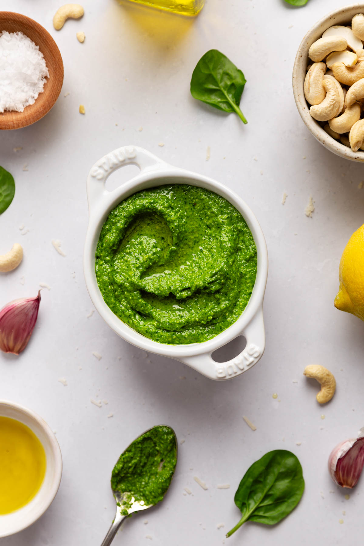 Top down image of spinach cashew pesto in a ceramic bowl on a light gray background with ingredients poking into the edges of the frame