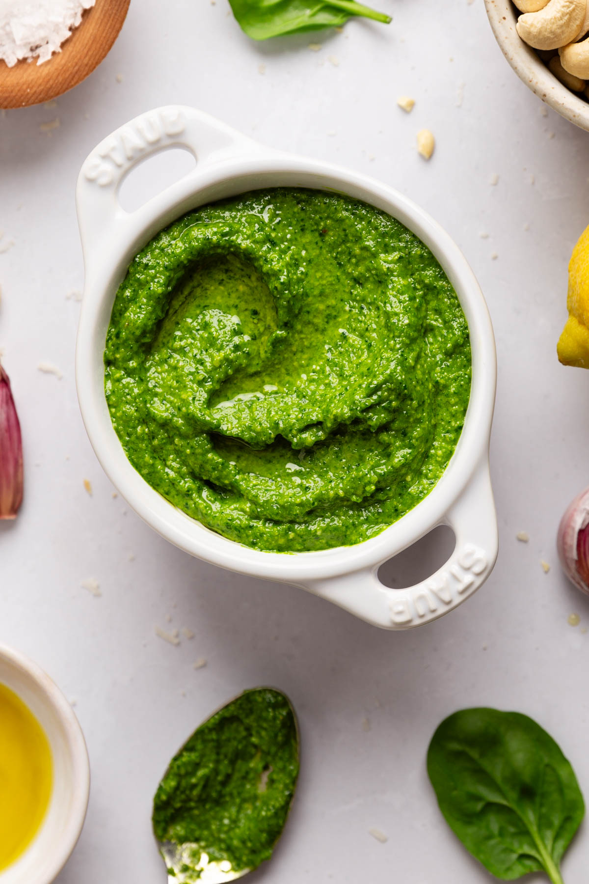 Beautiful top down photo of spinach cashew pesto in a ceramic white bowl with bright natural light coming in from the top of the frame with ingredients poking in around the sides