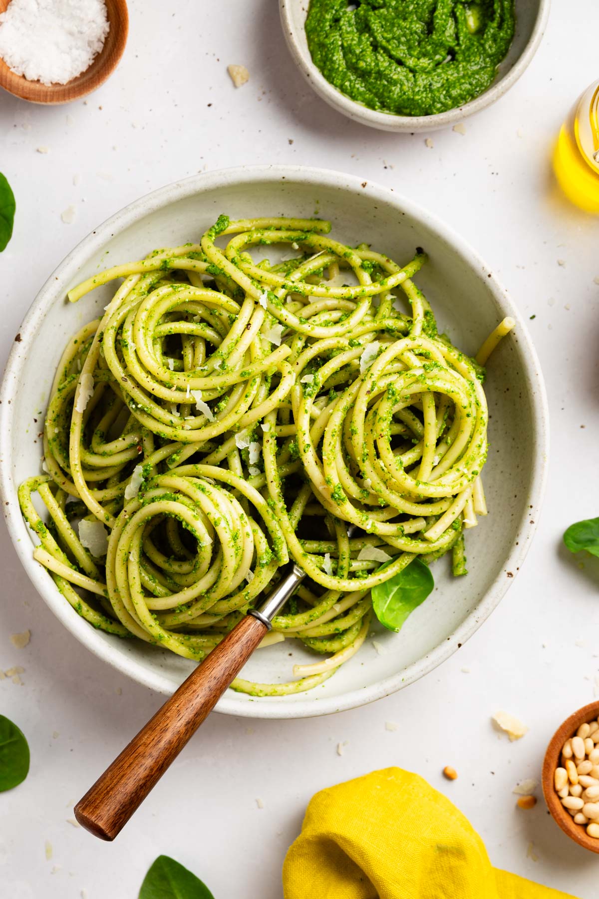 a medium overhead shot of a bowl of spinach pesto pasta with a wooden handle fork with ingredients poking in on the edges of the frame