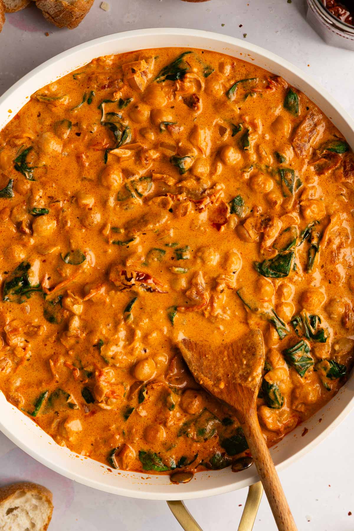 A closeup overhead photo of a skillet filled with creamy Tuscan chickpeas, also known as Marry Me Chickpeas, in a creamy sauce with sun-dried tomatoes, fresh wilted spinach and served with crusty bread.