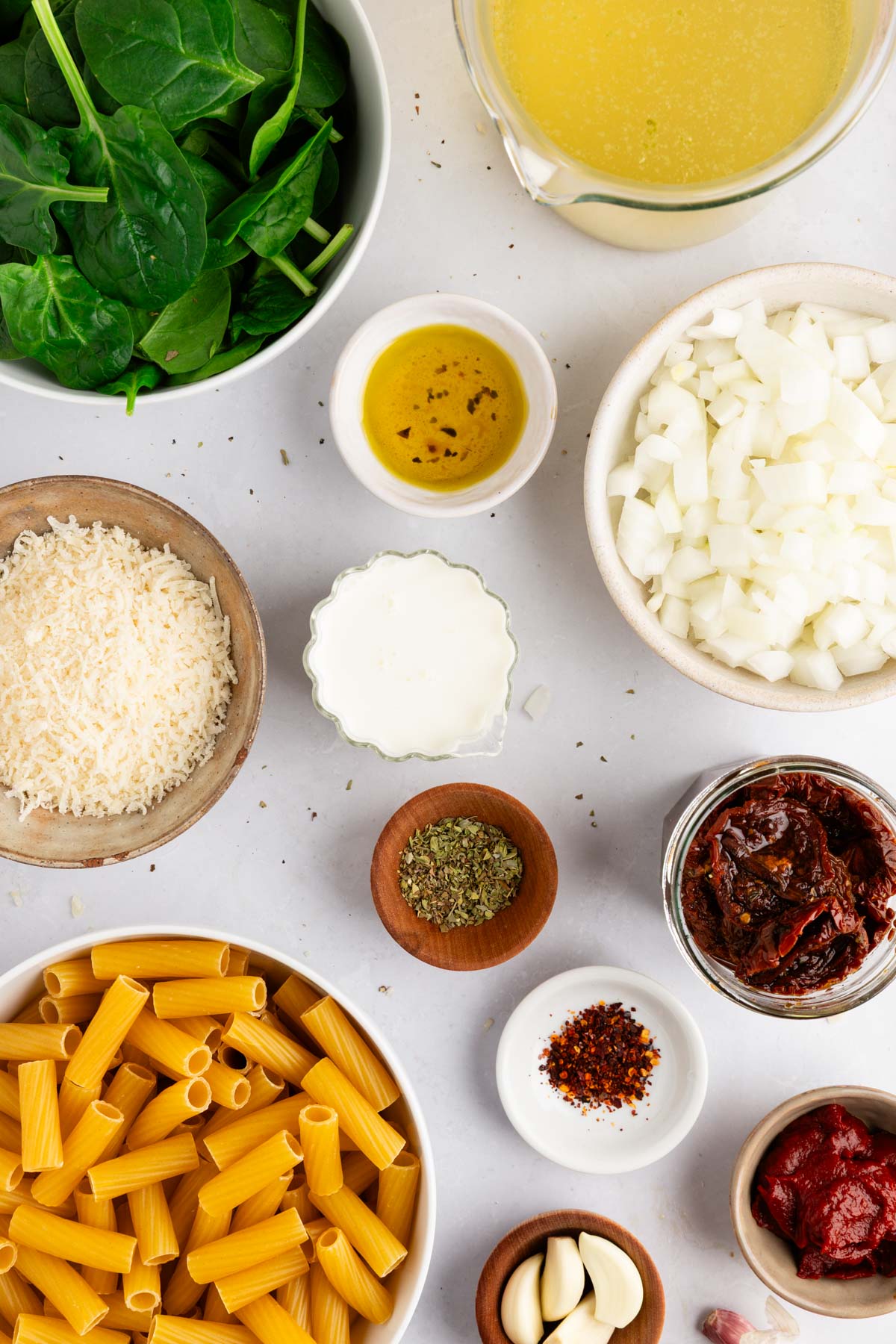 Flat lay of ingredients for Marry Me Pasta, including pasta, onion, garlic, sun-dried tomatoes, spinach, cream, Parmesan, and spices on a white surface.