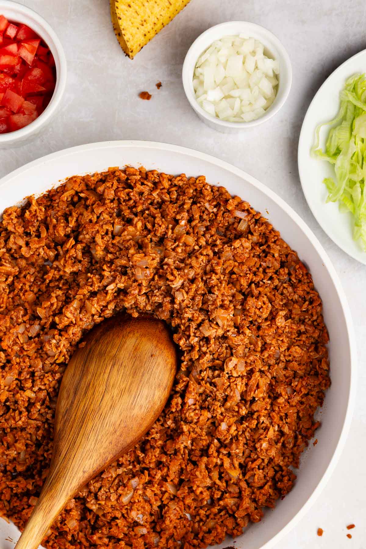 closeup of textured vegetable protein taco meat in a white skillet with a wooden spoon and bowls of taco fixings peeking into the frame