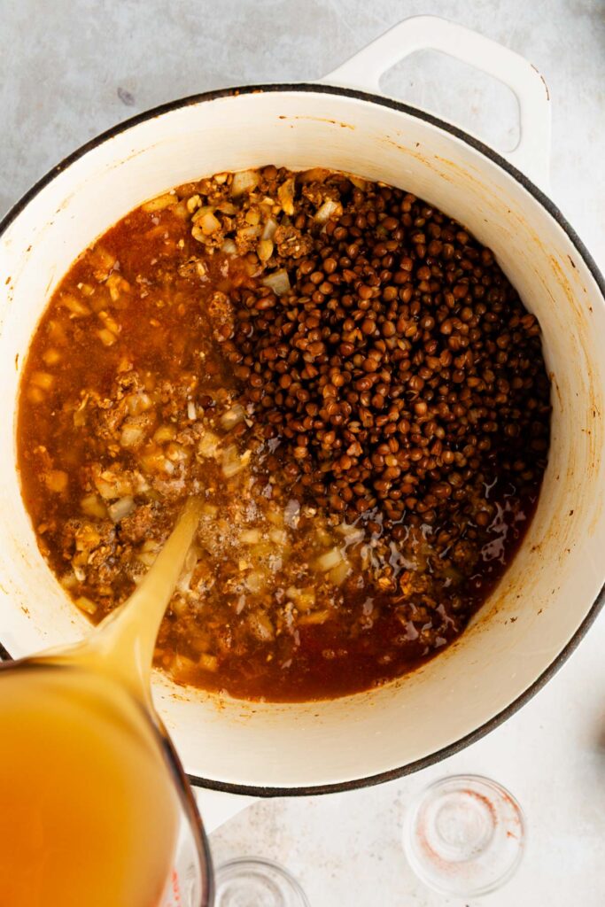 Pouring beef broth into a Dutch oven with the seasoned beef mixture and lentils.