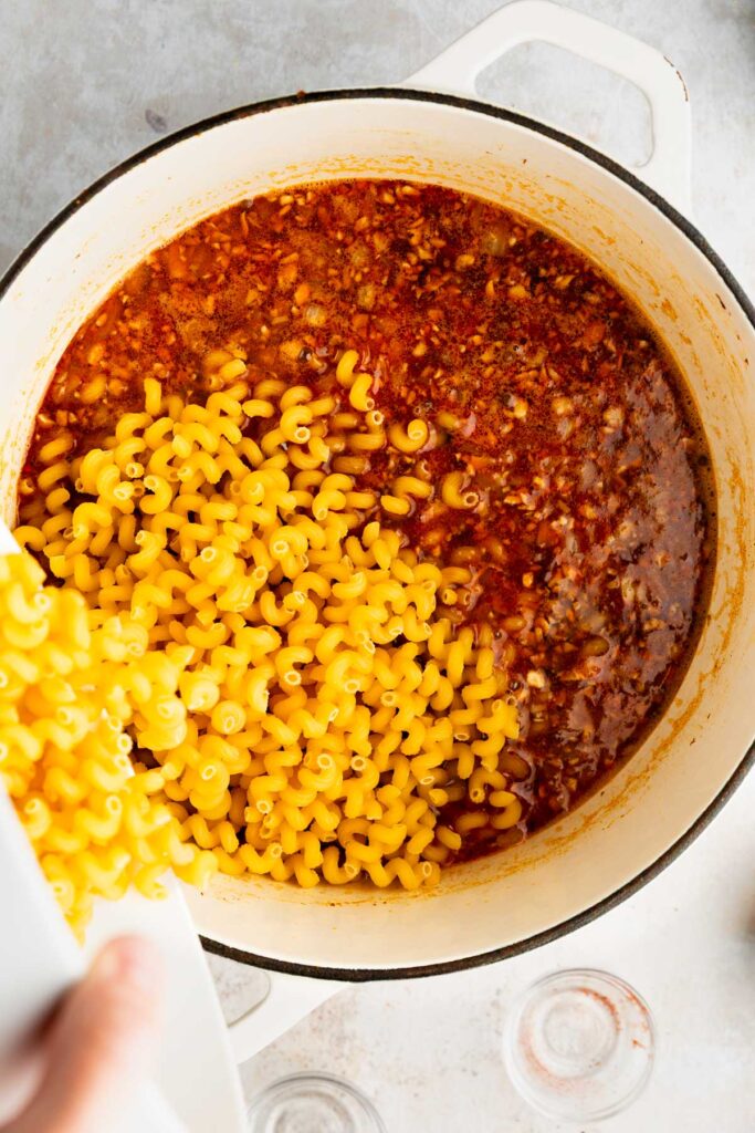 Pouring dry cavatappi pasta into broth and beef mixture in a Dutch oven.
