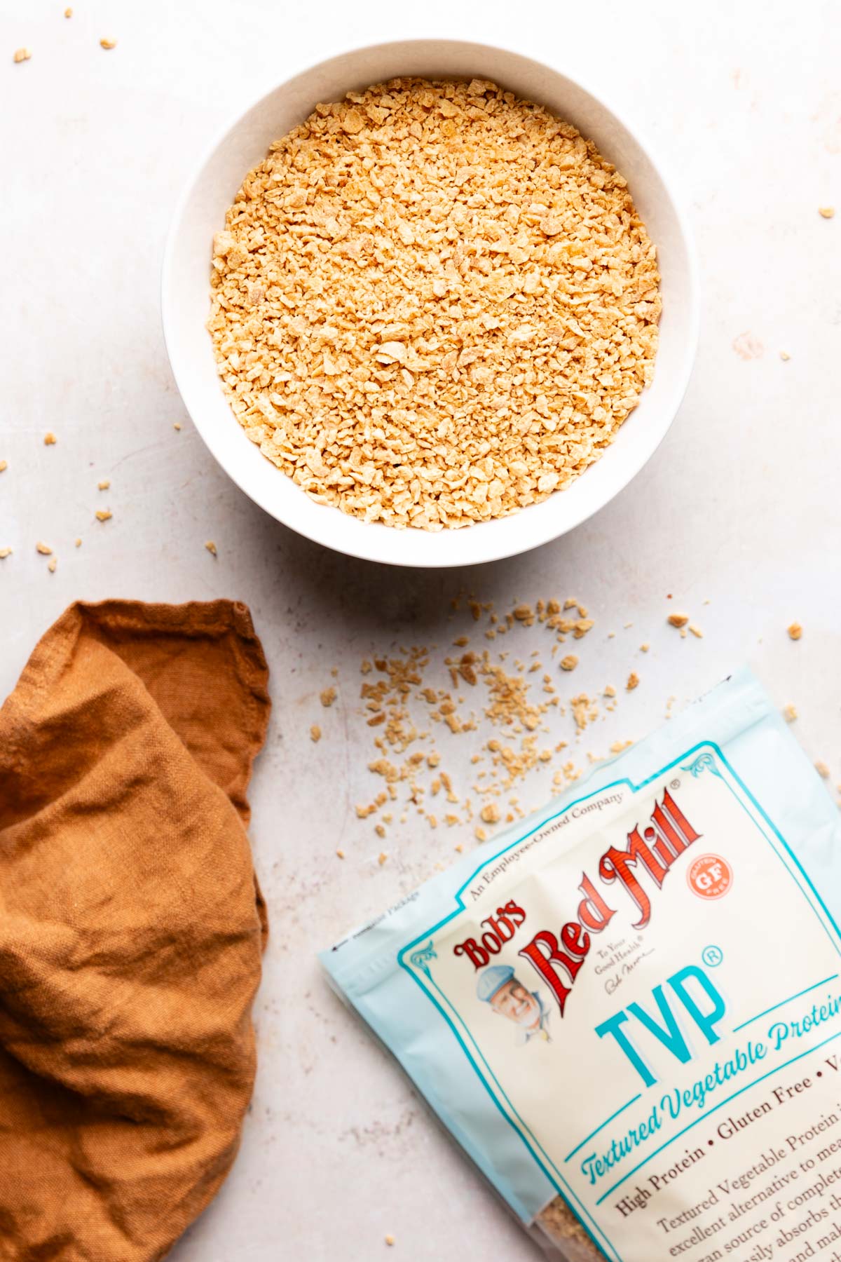 Dry textured vegetable protein (TVP) granules in a white bowl next to a bag of Bob’s Red Mill TVP and a rust-colored linen napkin.