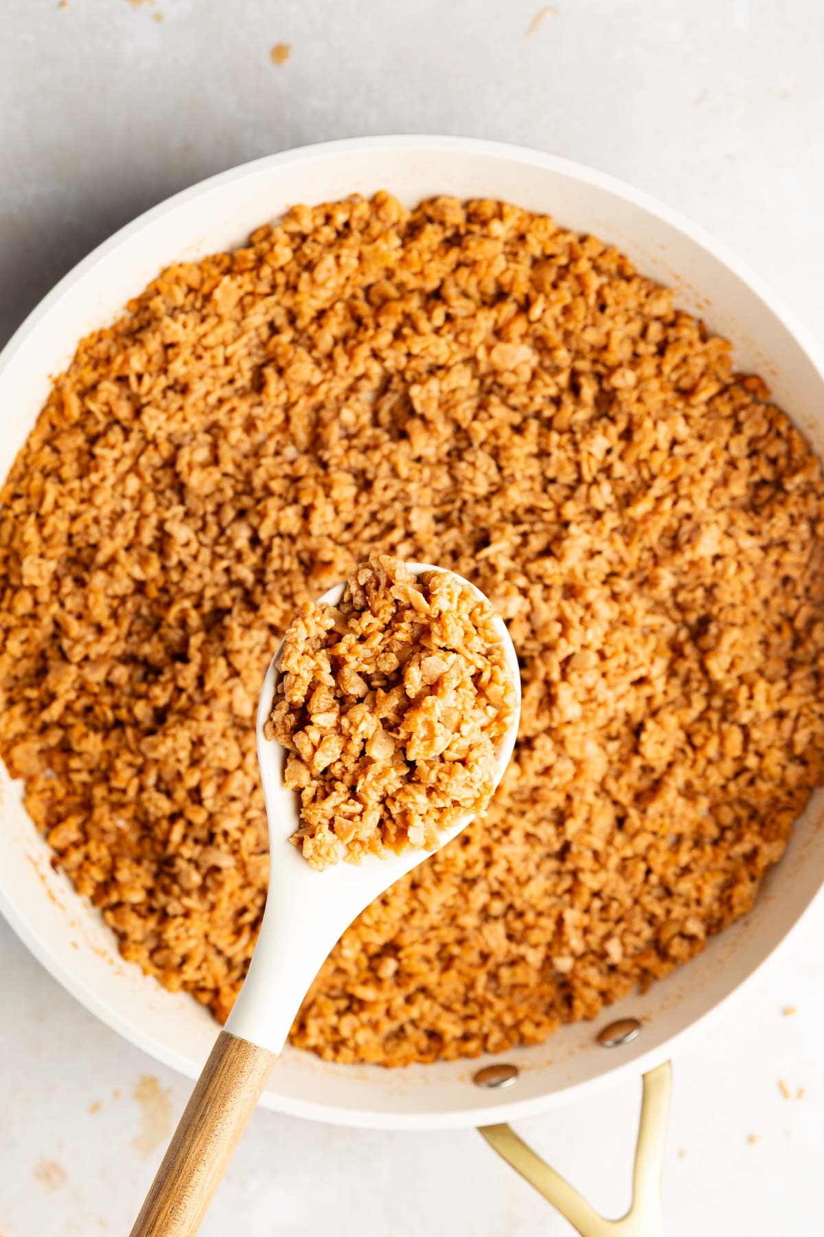 A white spoon lifting a scoop of TVP vegan ground meat from a skillet of cooked crumbles.