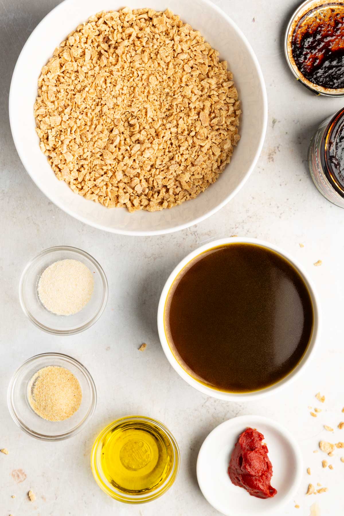 Ingredients for vegan ground meat in small bowls: dry TVP granules, vegan beef broth, garlic powder, onion powder, avocado oil and tomato paste.