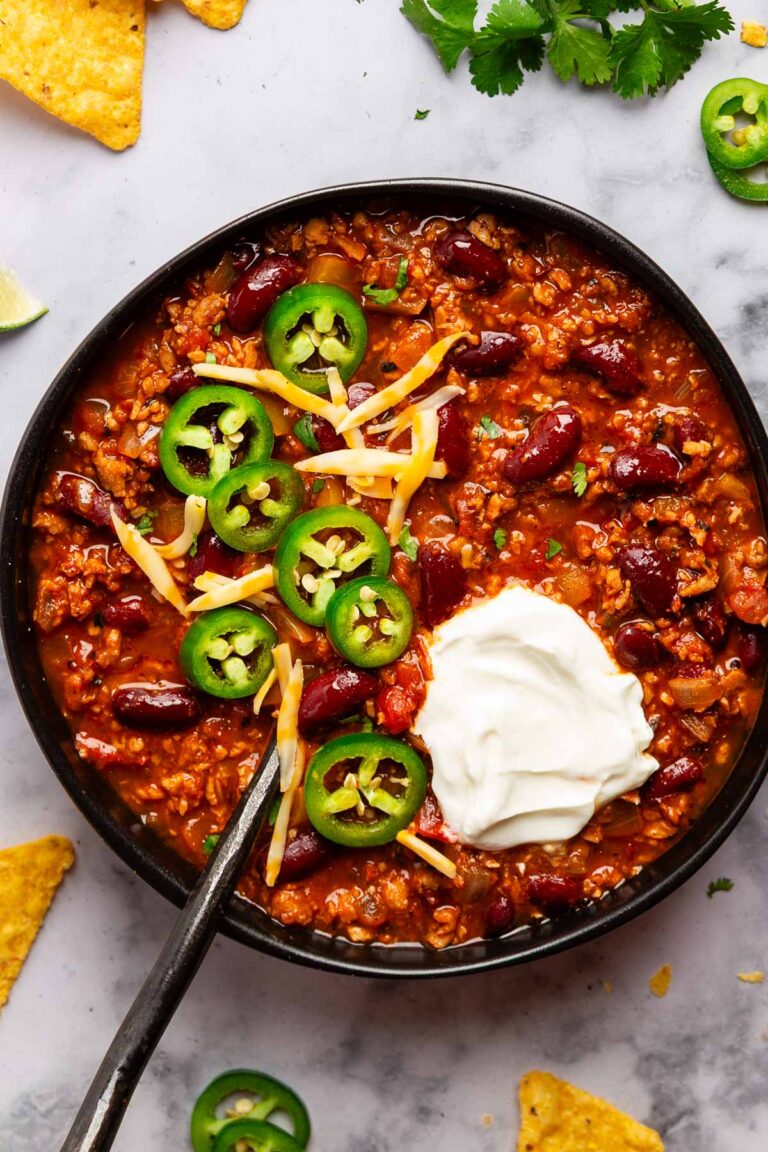 overhead photo of a black bowl with vegetarian chili with tvp topped with jalapenos, cheese, and sour cream on a marble background with ingredients and chips poking into the frame