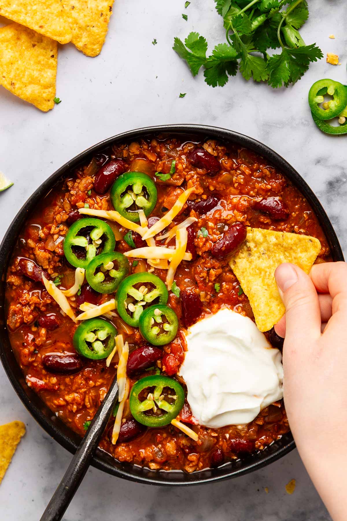 overhead photo of a hand scooping vegetarian tvp chili with a tortilla chip from a bowl topped with jalapenos, cheese, and sour cream