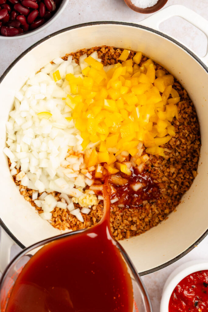 Overhead photo of onions, pepper,s and broth being added to a pot of tvp for chili