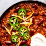 Overhead view of a black bowl of vegetarian chili topped with jalapeño slices, shredded cheese and a dollop of sour cream, with tortilla chips and cilantro nearby. Text at the top reads Kid-Approved Vegetarian Chili, mild flavor, one pot, easy and healthy.